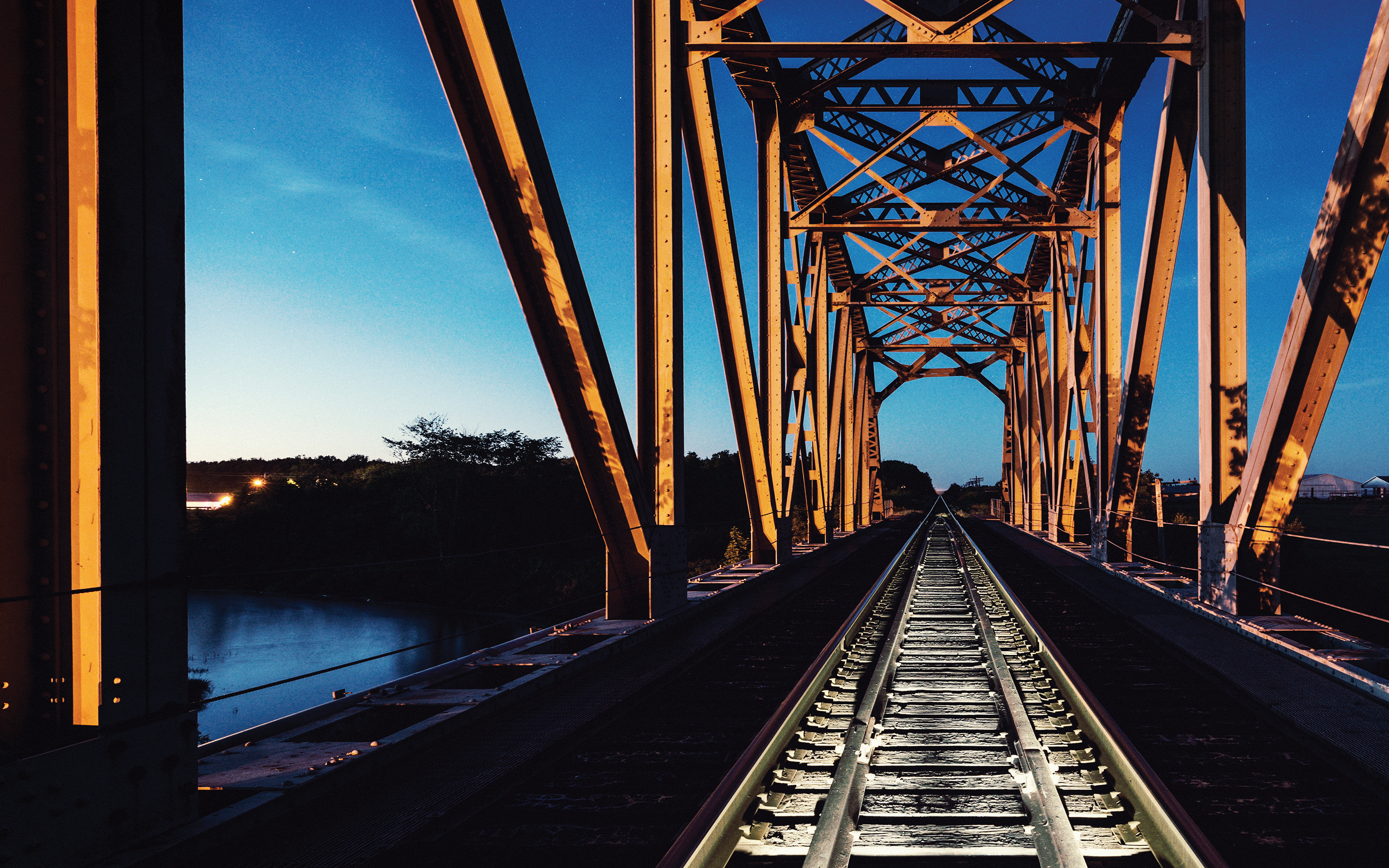 Railroad tracks running through an orange steel truss bridge at dusk, with a river and tree line visible to the side.