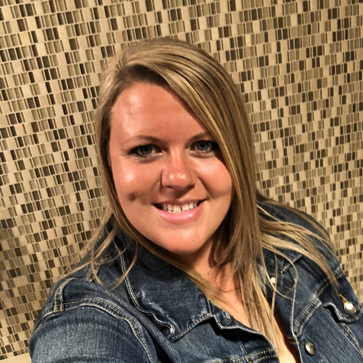 Smiling woman with long blonde hair wearing a denim jacket, sitting in front of a patterned wall.