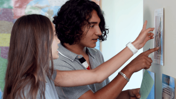 Two students point at and discuss information on a paper posted to the wall in a classroom setting.