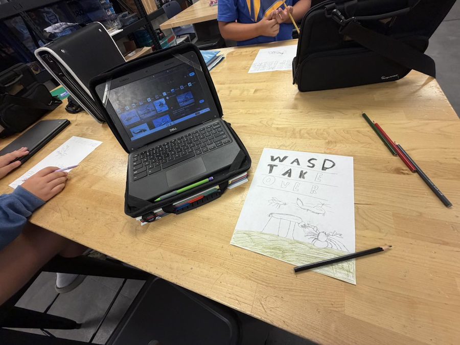 A student draws WASD TAKE OVER on paper beside a laptop and colored pencils on a classroom table.