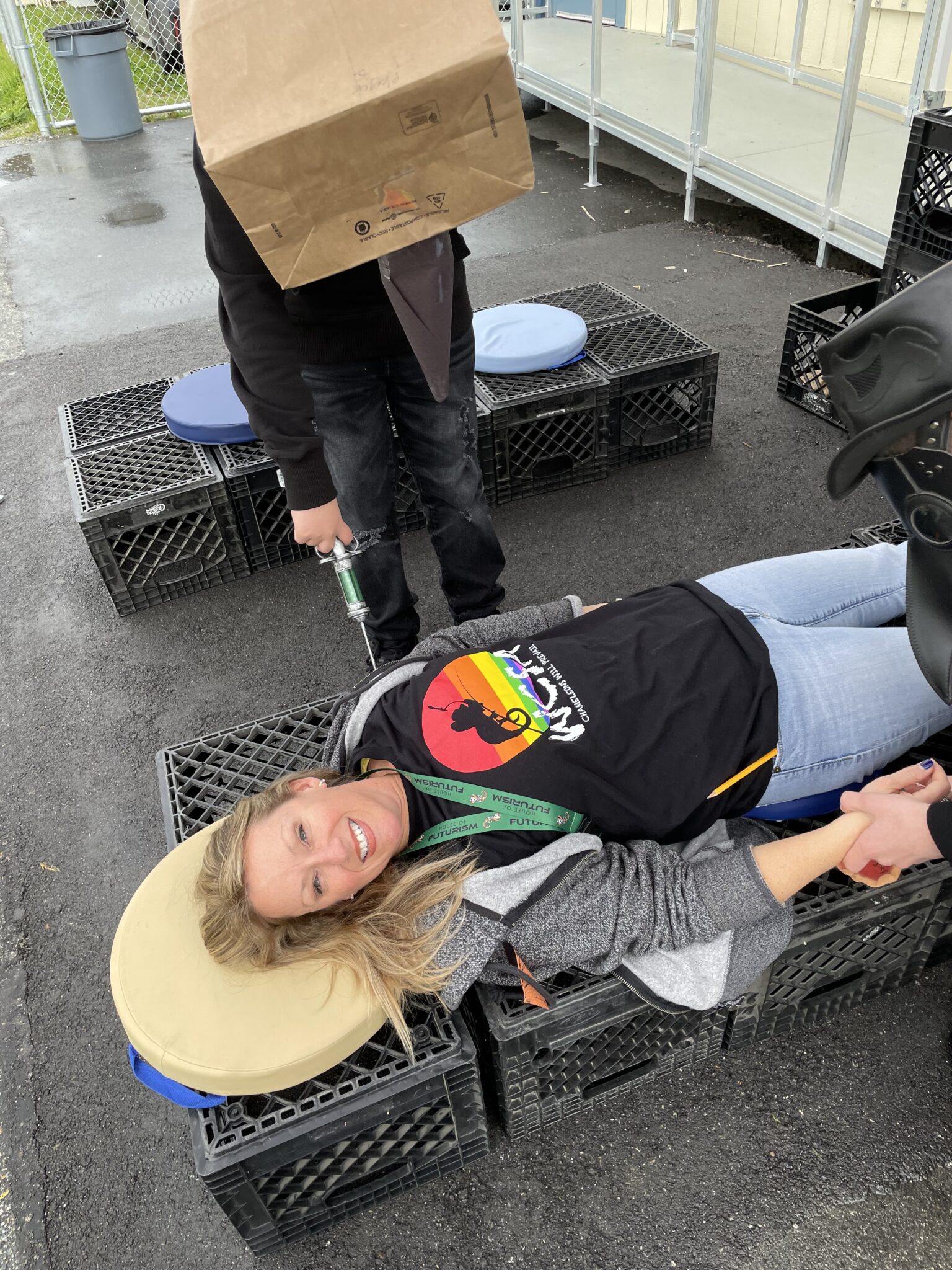 A smiling woman lies on crates while another person stands nearby with a paper bag over their head.