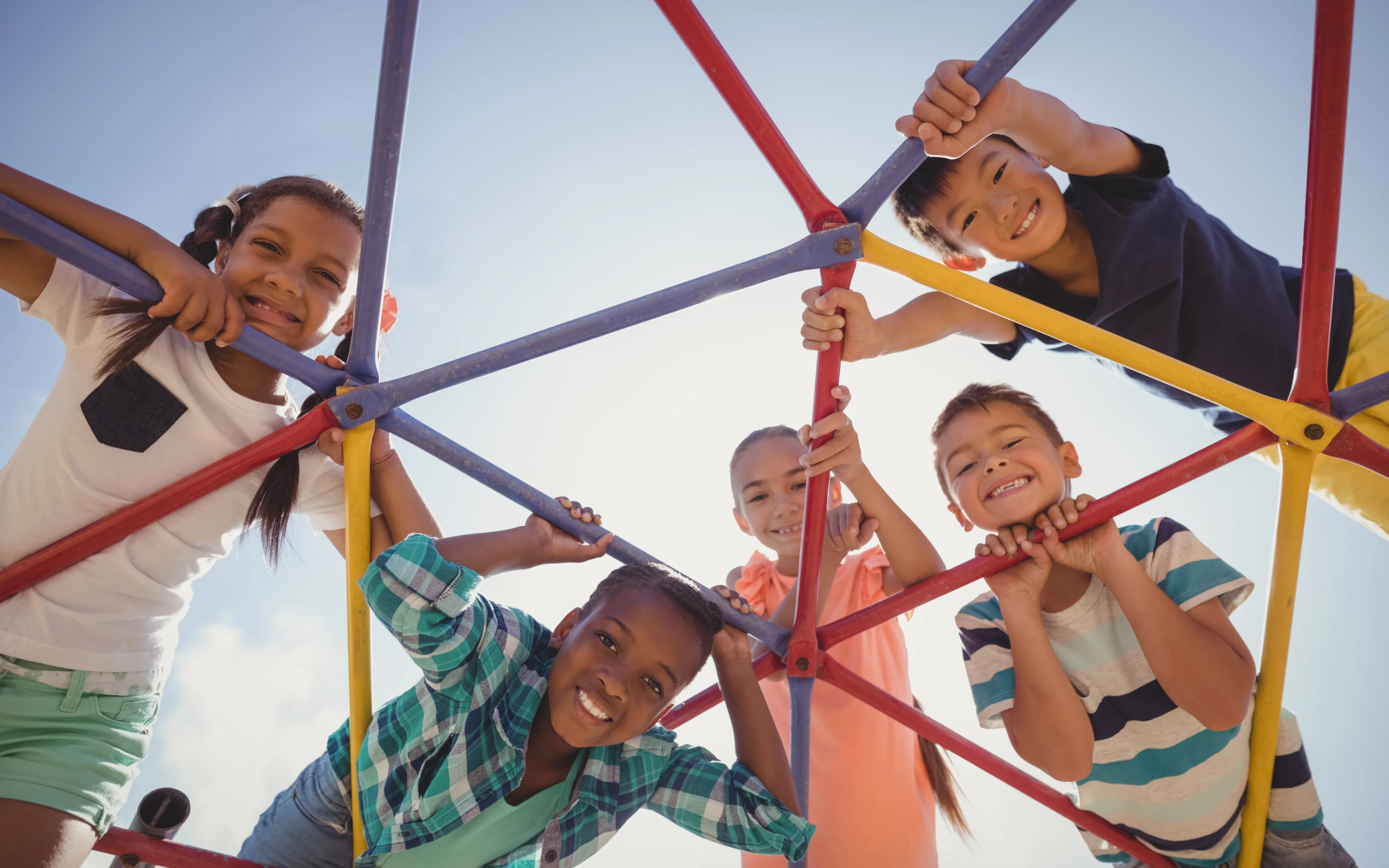 Five smiling children climb and play on a colorful jungle gym, looking down at the camera on a sunny day.