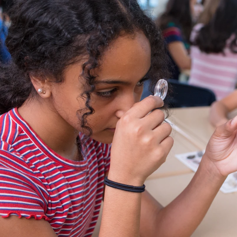 Girl in a red striped shirt examines something closely with a magnifying glass at a classroom table.