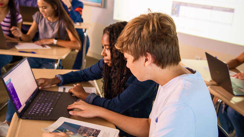 Two students work together on a laptop in a classroom, surrounded by classmates at their desks.
