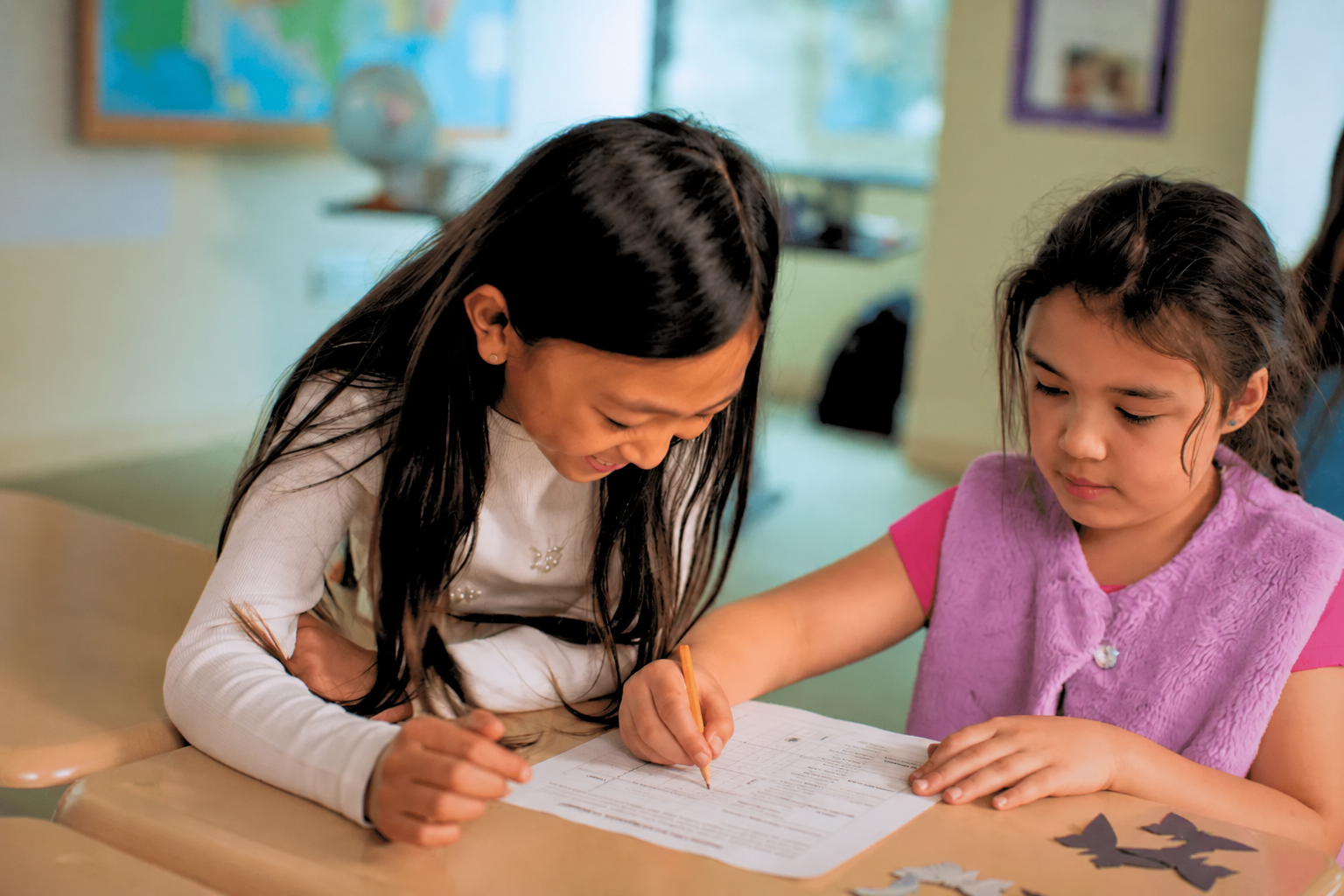 Two girls sit at a desk, smiling and working together on a worksheet in a classroom setting.