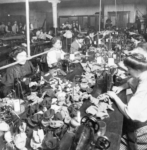 Women work at sewing machines in a crowded factory, assembling fabric items, with supervisors in the background.