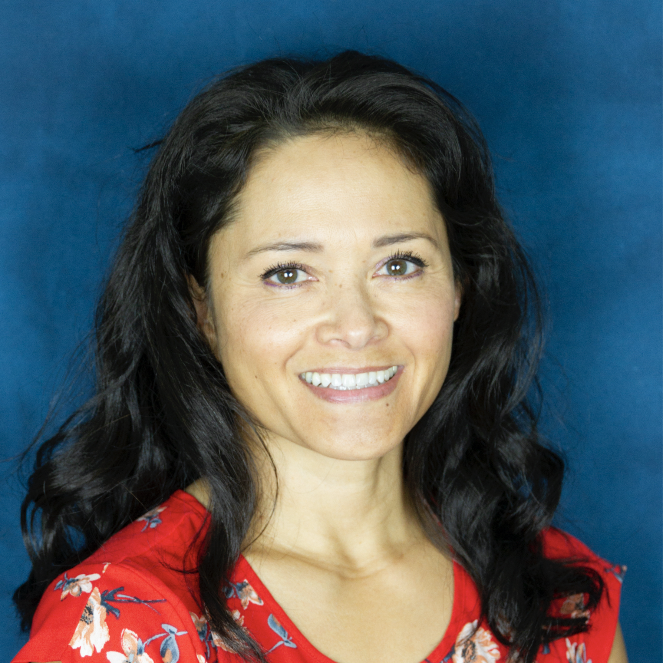 Woman with long dark hair and a red floral top smiling in front of a blue background.
