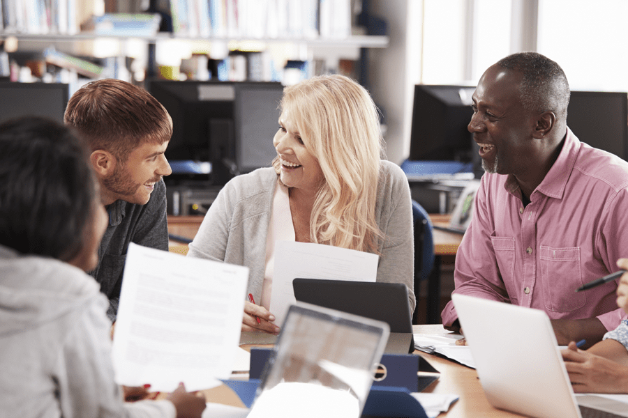 Four adults sit at a table in an office, smiling and talking, with papers and laptops in front of them.
