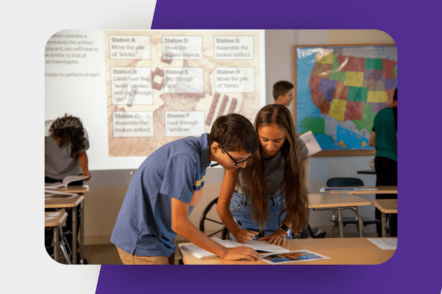 Two students work together at a desk in a classroom while a projected slide details archaeology-themed stations.