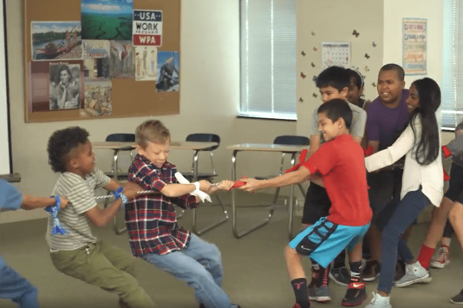 Children play tug-of-war in a classroom, pulling a rope and smiling, with desks and posters in the background.