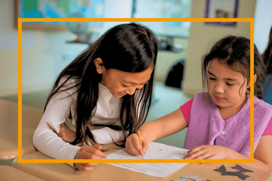 Two young girls sit at a desk, working together on a worksheet in a classroom setting.