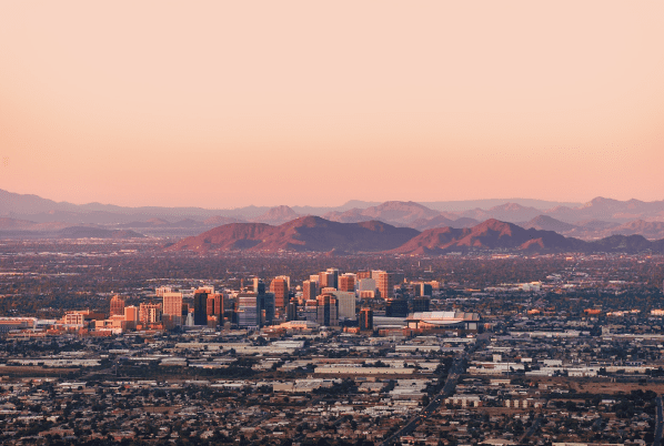 Downtown cityscape with tall buildings, surrounded by desert, mountains, and a soft pink sky at sunset.
