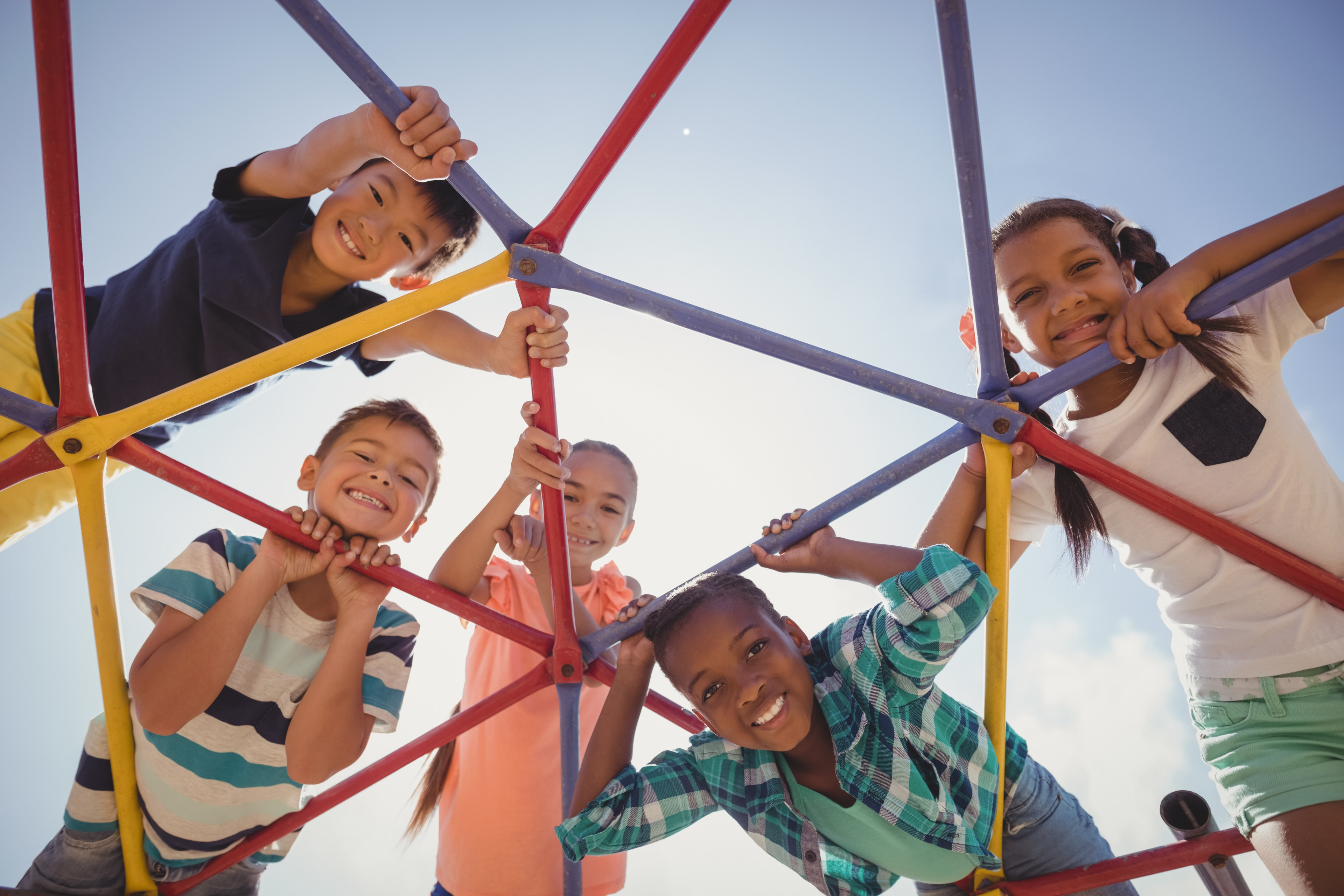 Five smiling children look down through playground bars, framed by blue sky, enjoying a sunny day outside together.