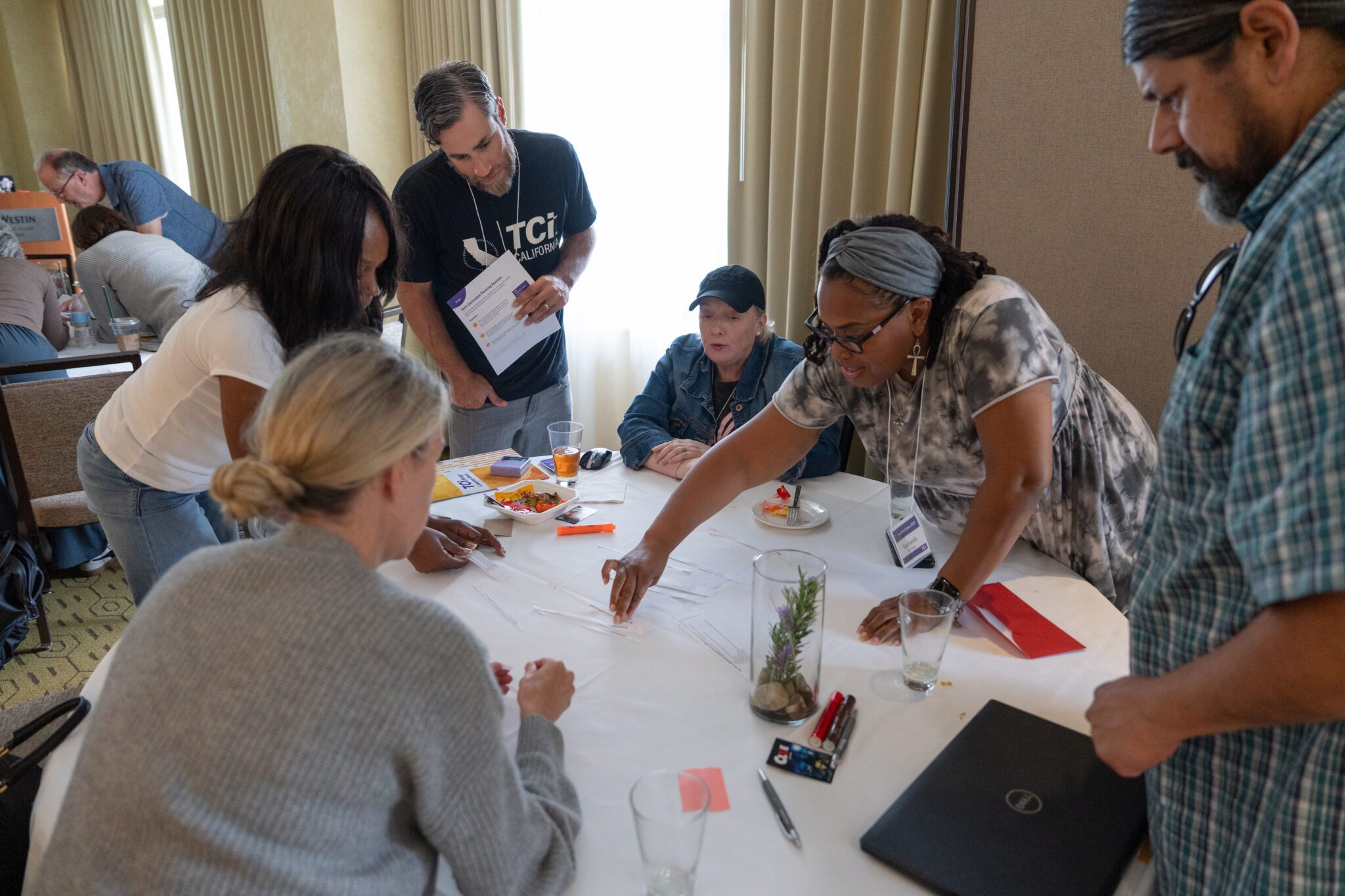 Six adults gather around a table, collaborating over papers, snacks, and drinks in a meeting room.
