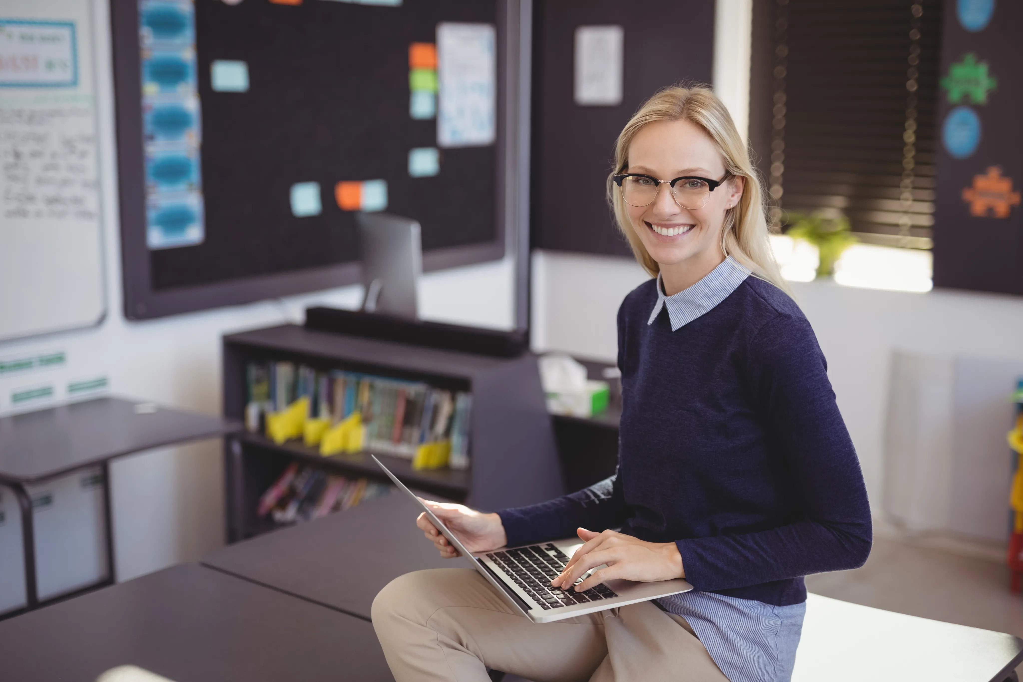 Smiling woman wearing glasses sits on a desk with a laptop in a classroom setting.