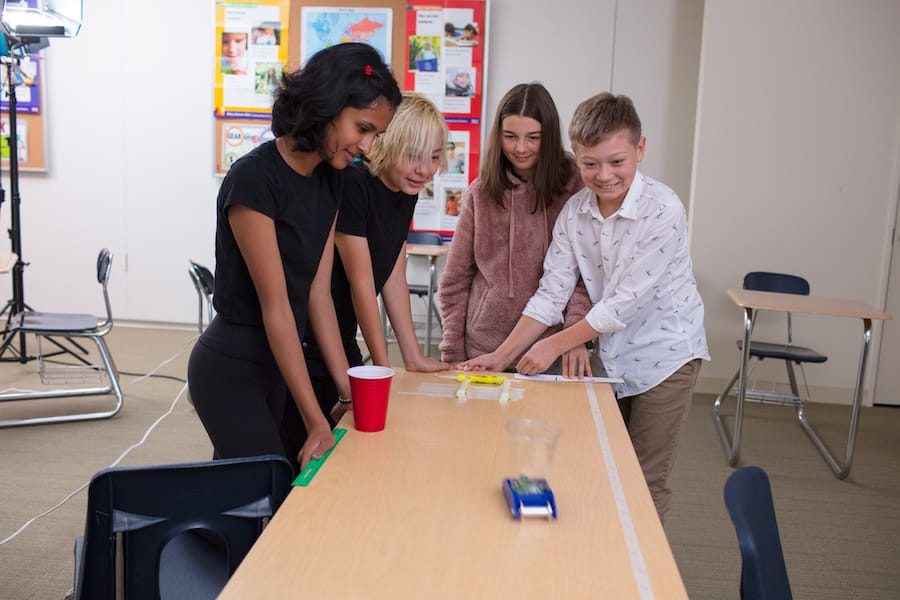 Four kids in a classroom watch a small toy car move across a table, smiling and engaged in the activity.