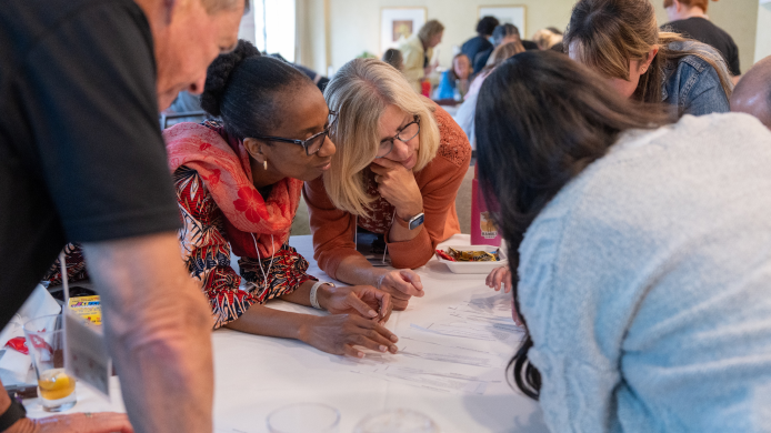 A diverse group of people lean over a table, collaborating and discussing papers during a meeting.