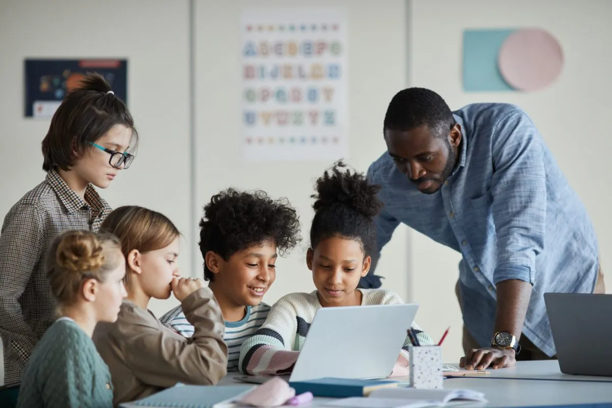 A teacher helps a diverse group of students working together on a laptop in a classroom.