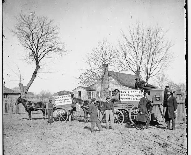 Black-and-white photo of men with two horse-drawn wagons in front of a house and leafless trees.