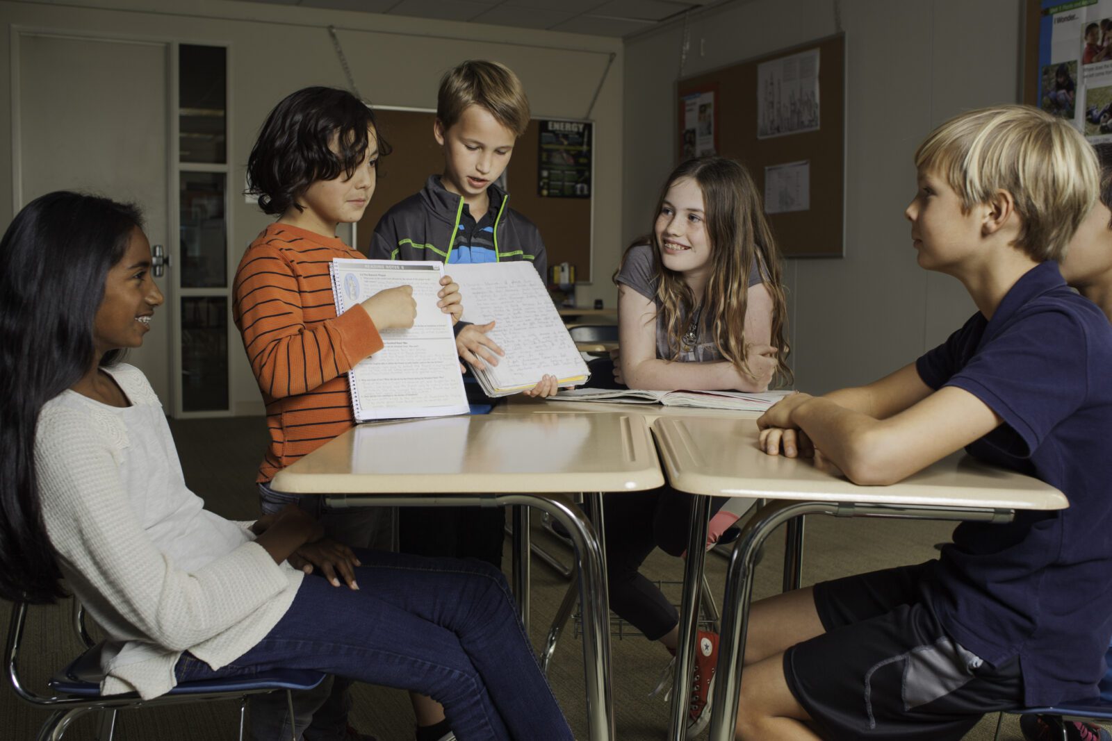 Five students sit at desks, sharing notebooks and discussing together in a classroom setting.