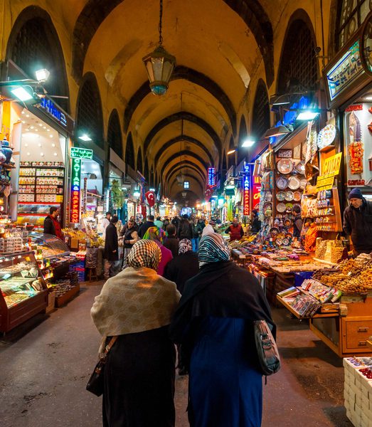 People walk through a vibrant indoor market with colorful lights, food stalls, and arched ceilings.
