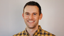 A man with short hair smiling, wearing a yellow and black plaid shirt.