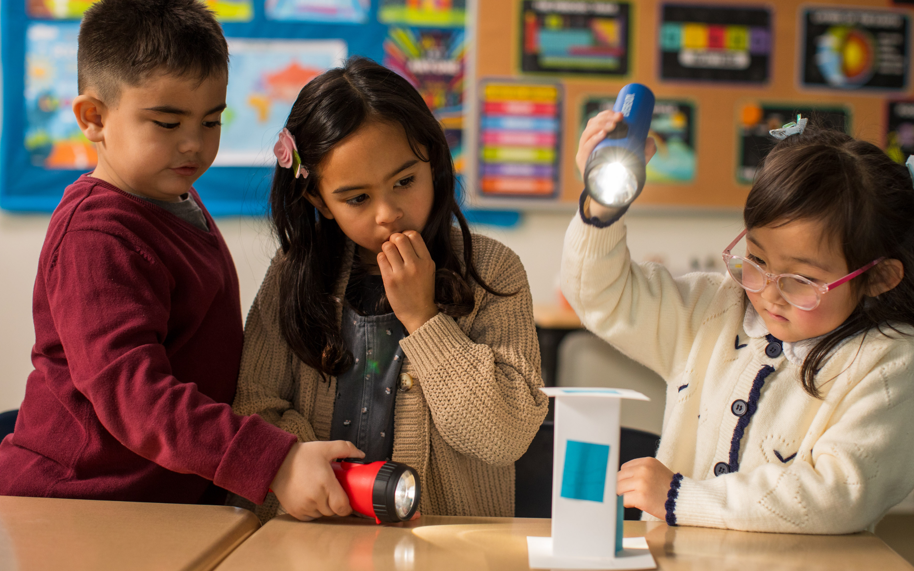 Three children shine flashlights on a paper structure in a colorful classroom, observing shadows and light.