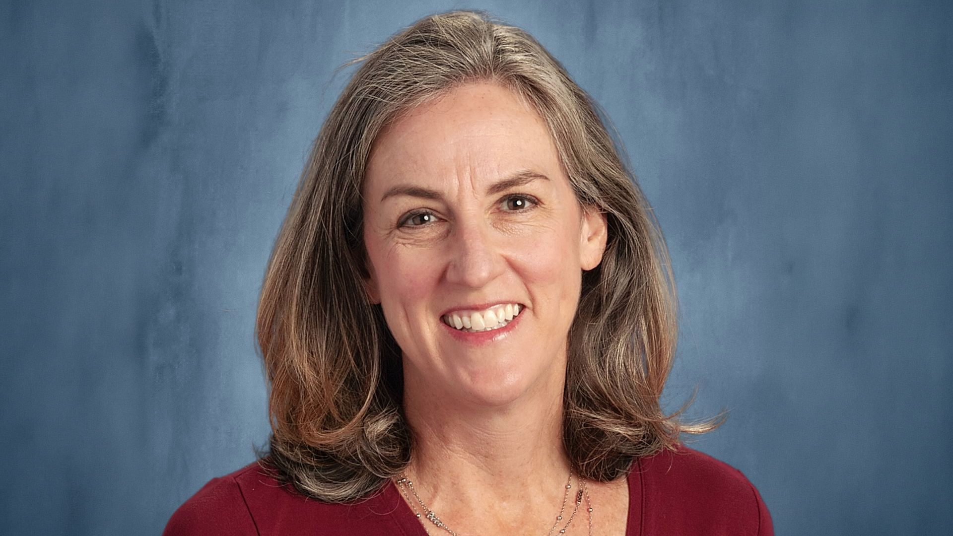 Smiling woman with shoulder-length brown hair wearing a maroon top, posed in front of a blue background.