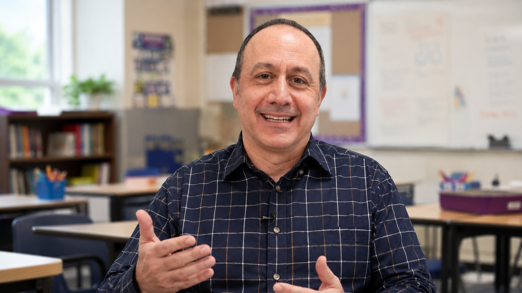 A man in a checkered shirt smiles and gestures while sitting in a brightly lit classroom.
