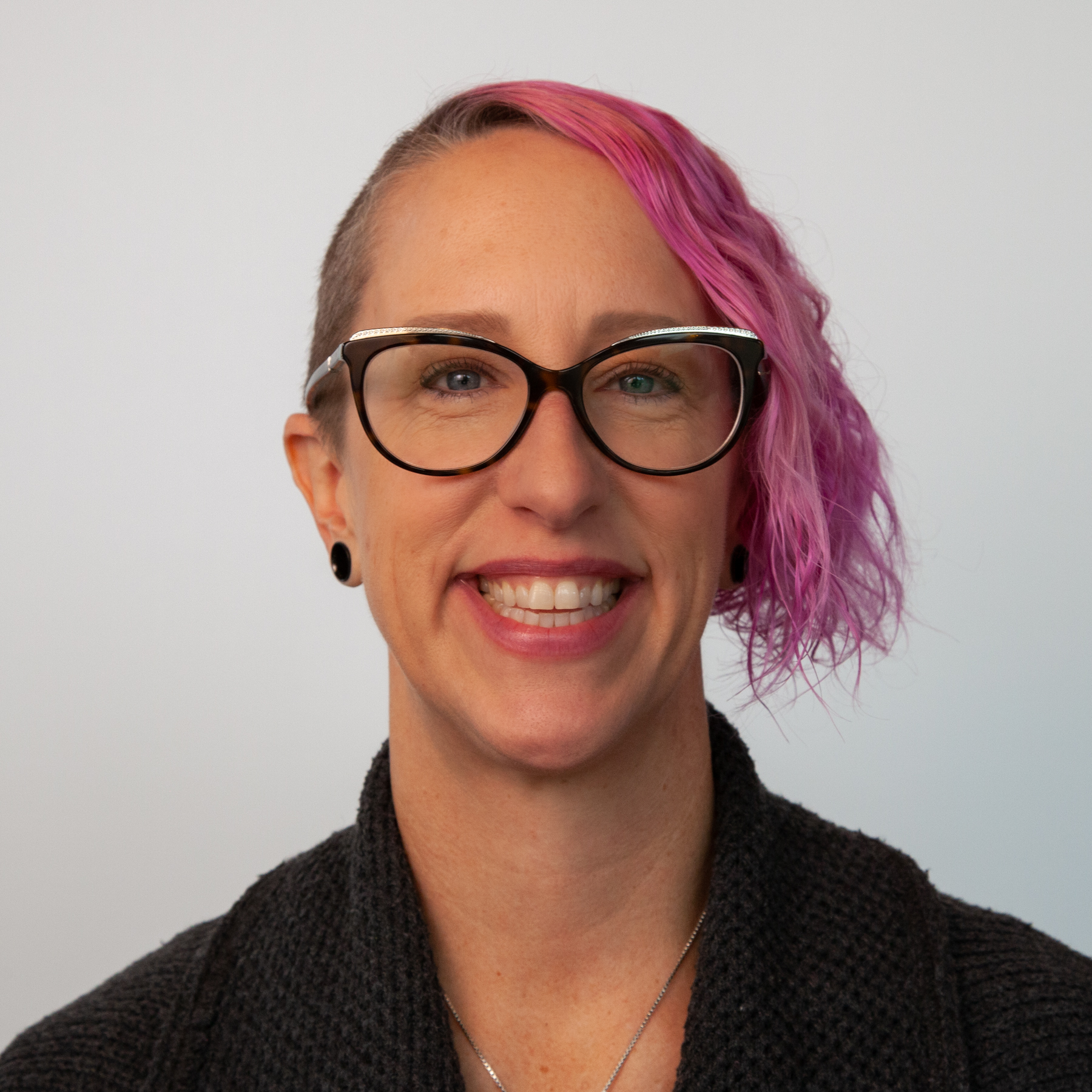 Smiling person with glasses, short pink hair on one side, and a dark sweater against a plain background.