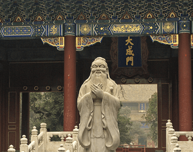 Stone statue of a bearded figure stands before an ornate traditional Chinese temple gate with red columns.