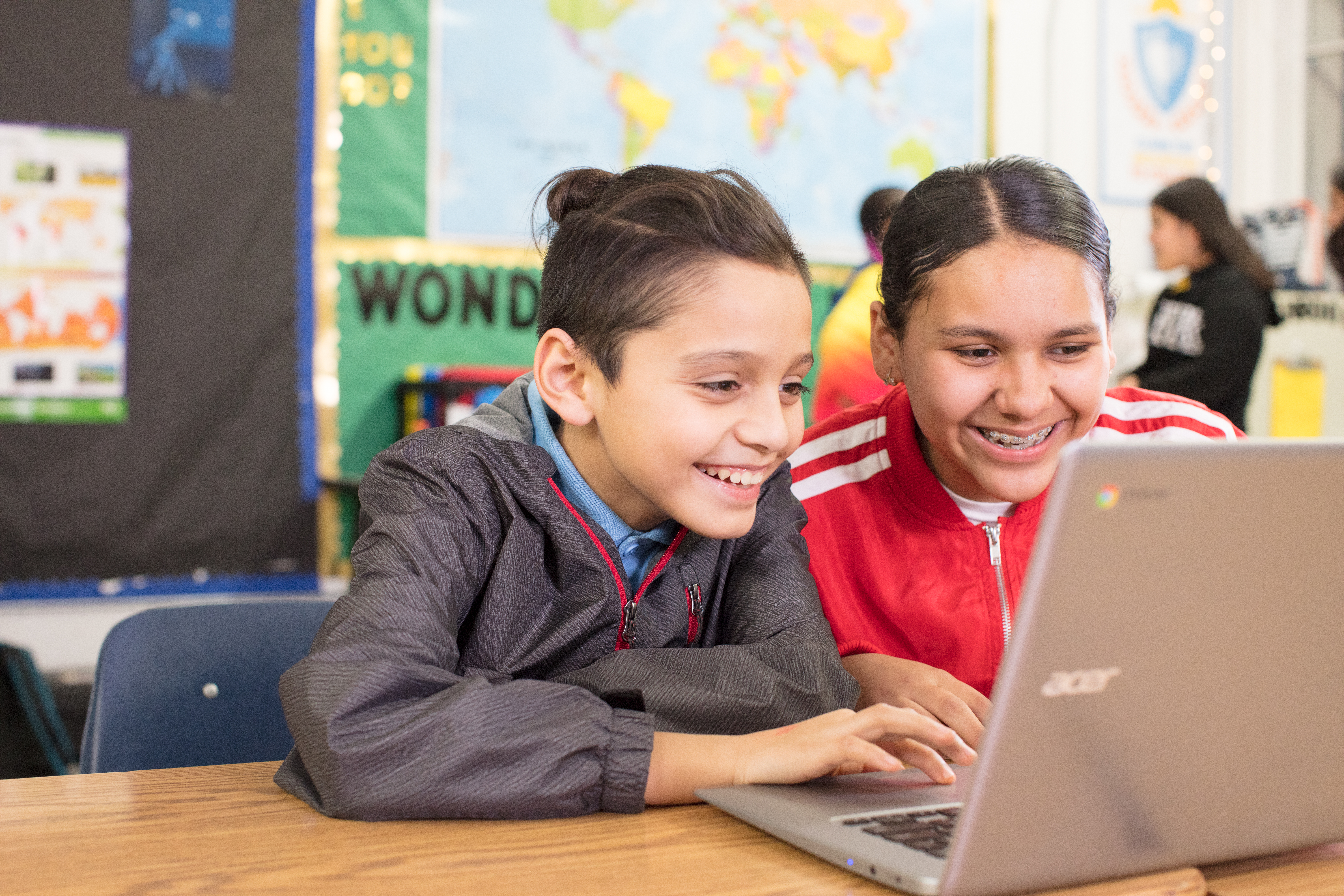 Two smiling middle school students sharing a Chromebook in a classroom with a world map and bulletin boards behind them.