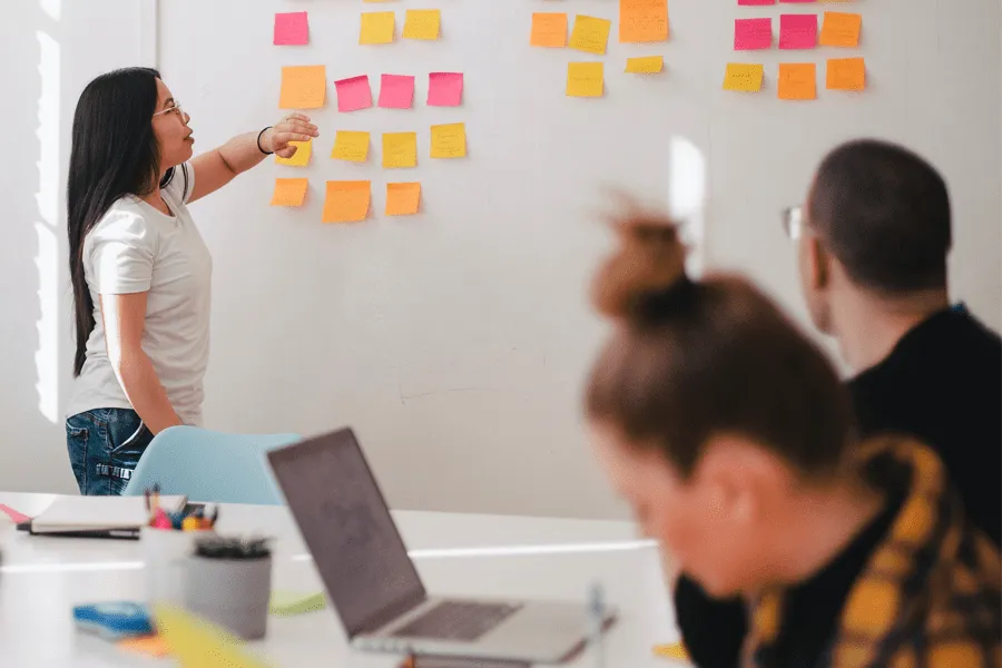 Woman points at sticky notes on a wall during a meeting while two people sit at a table with a laptop.