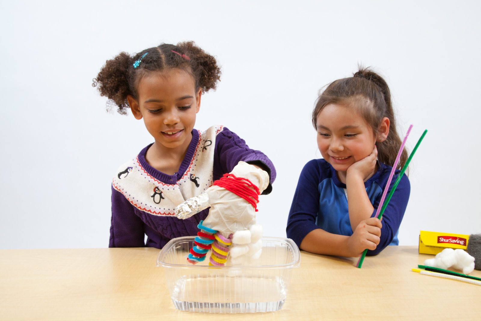 Two girls do a science experiment with a cotton and pipe cleaner figure over a tray of water at a table.