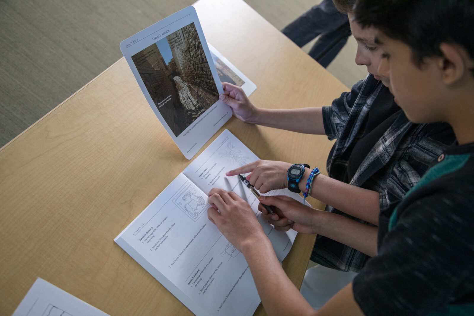 Two students work together on an assignment, looking at a worksheet and a printed photo at a classroom desk.