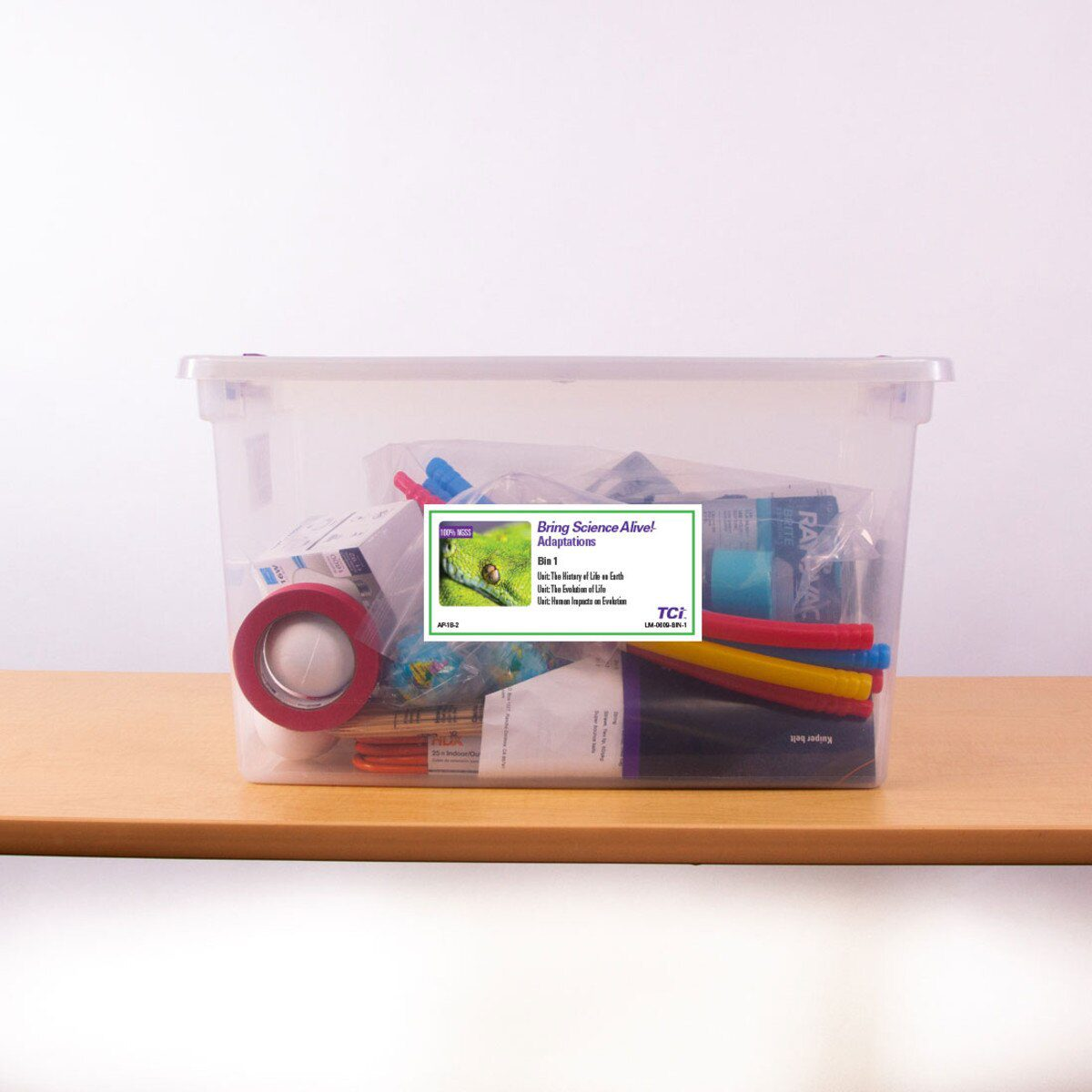 Clear plastic bin filled with school supplies like tape, markers, and colored paper, on a wooden surface.