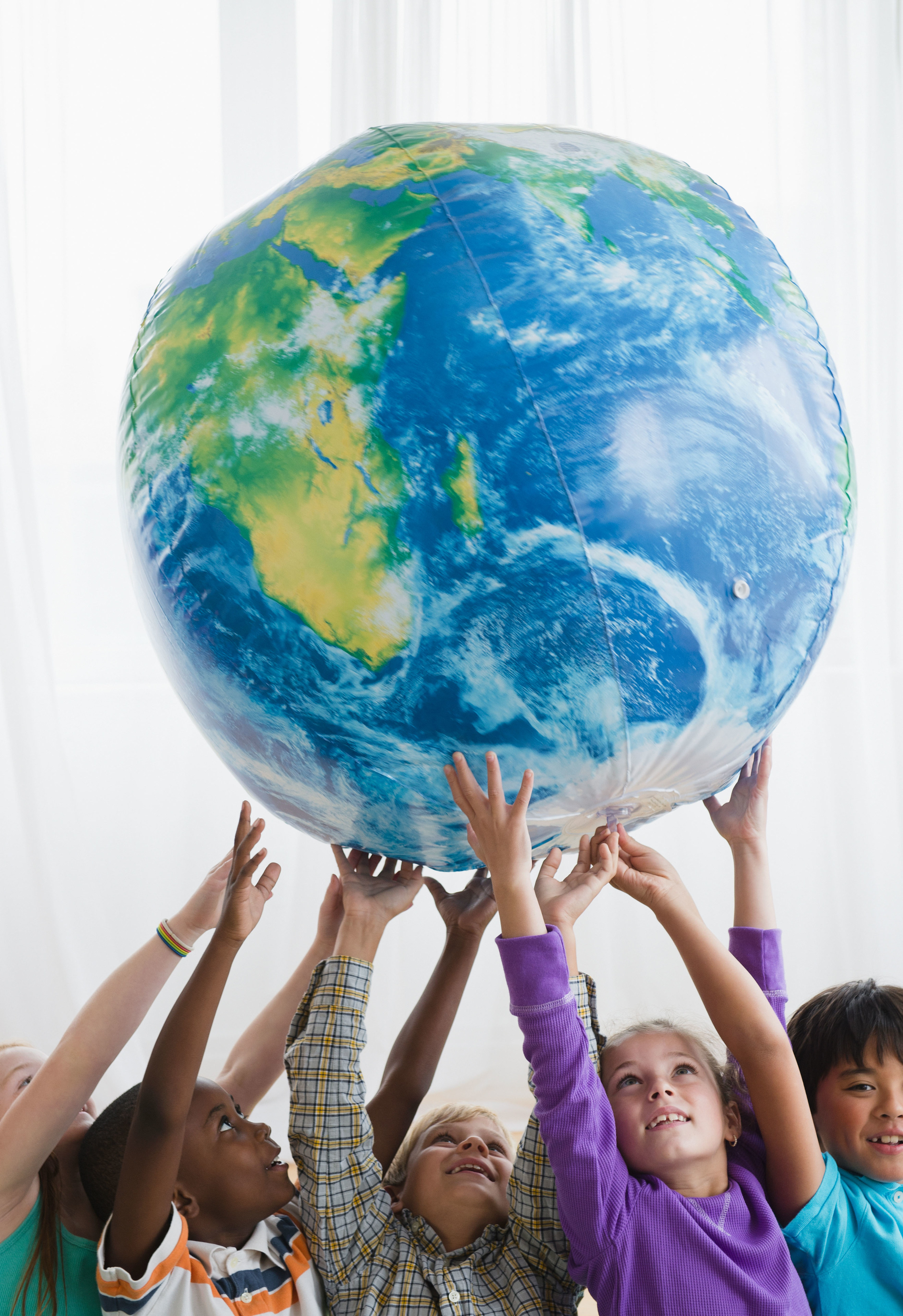 Children smiling and reaching up to hold a large inflatable globe together indoors.