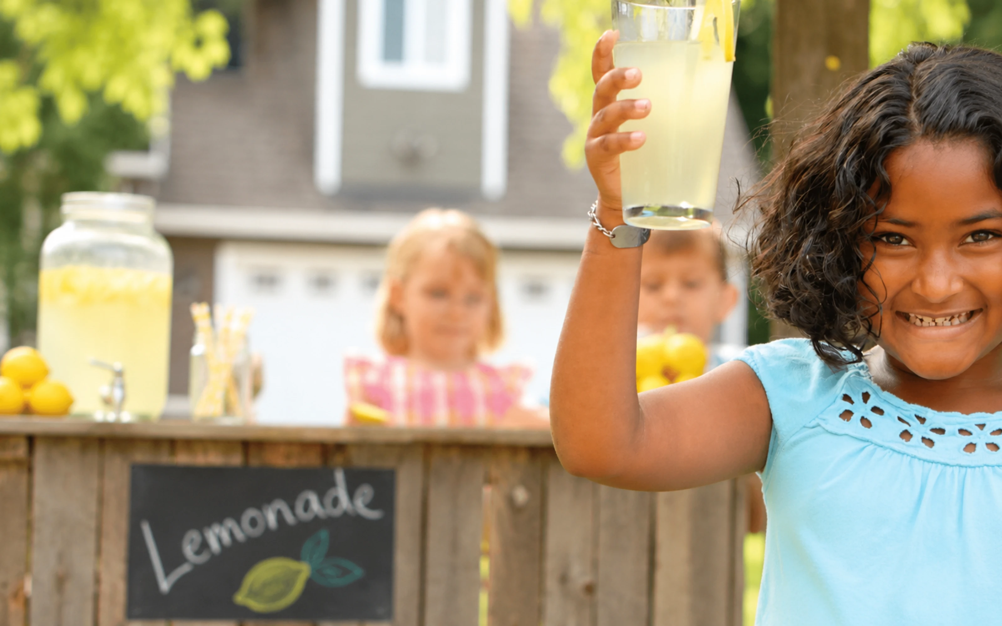 Smiling child holds up lemonade at a stand, with two kids and a Lemonade sign in the background.