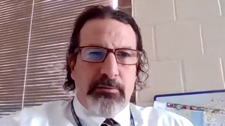 Man with glasses, dark hair, and a goatee wearing a white shirt sits indoors near a window and a corkboard.