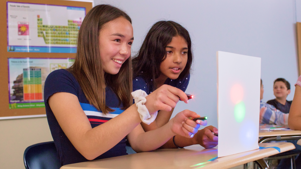 Two girls in a classroom shine colored lights on a white board, mixing colors. Other students sit in the background.