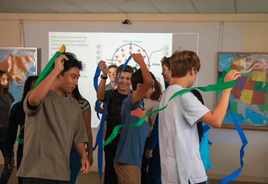 Students holding colorful streamers celebrate in a classroom with maps and a projected hurricane diagram behind them.