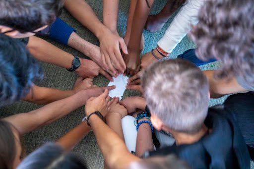 A group of people sit in a circle, reaching their hands toward a piece of paper in the center.