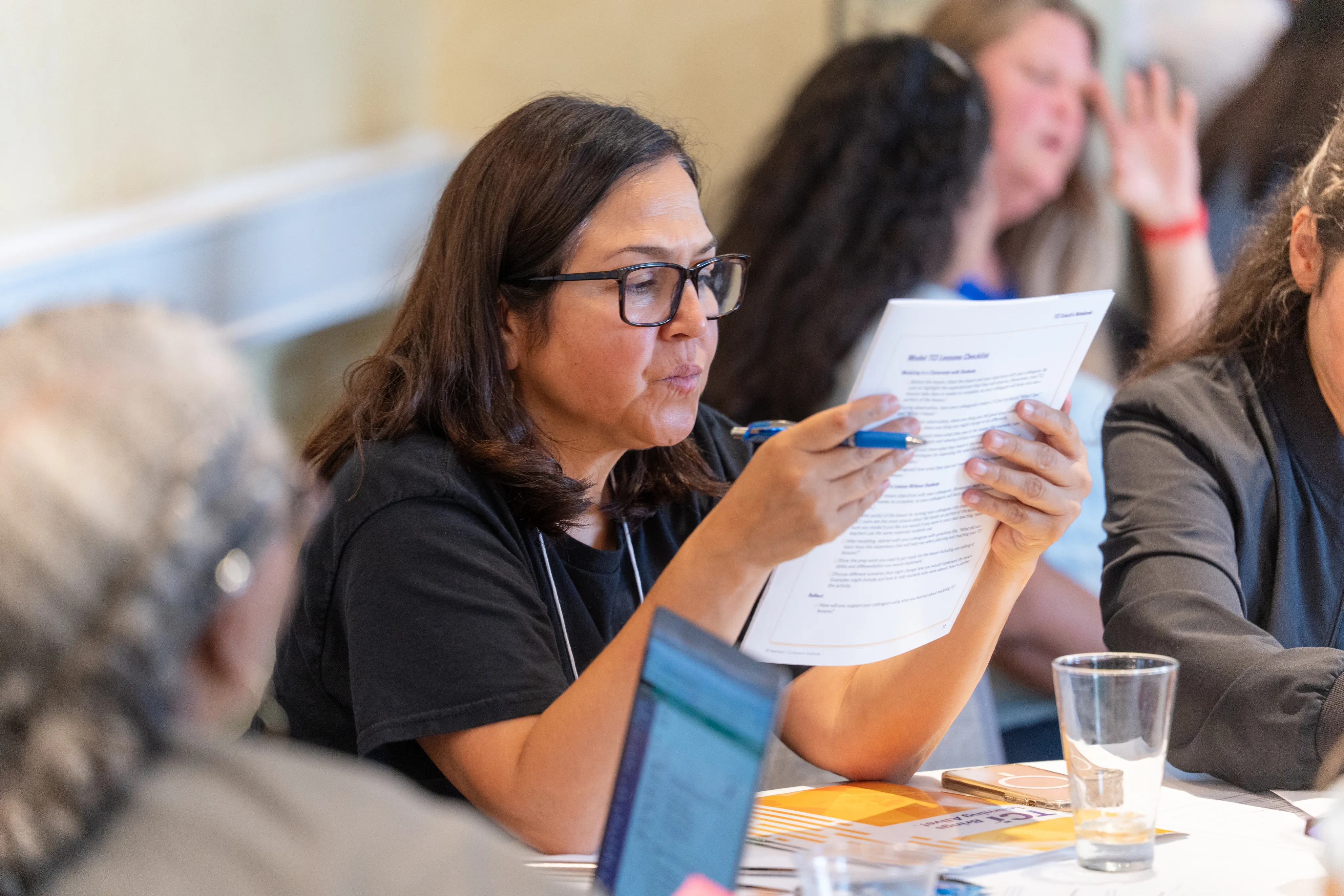 Woman with glasses reads a printed document at a table during a group meeting or workshop.