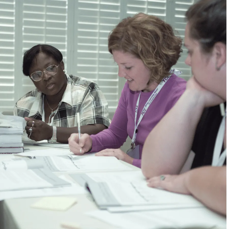 Three women sit at a table, writing on papers and discussing documents in a brightly lit room with white shutters.