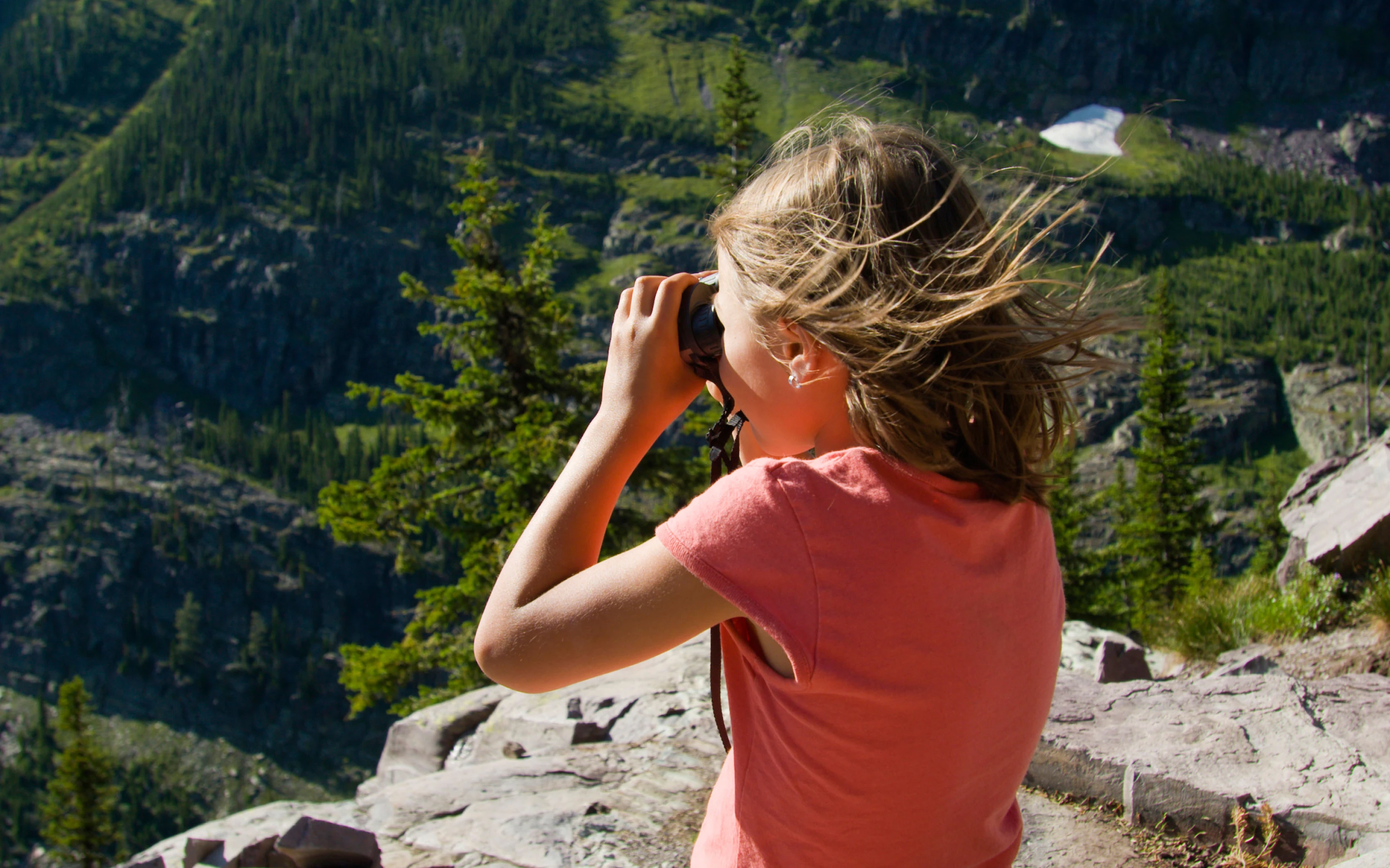 Girl with windswept hair uses binoculars while standing on a rocky mountain ledge overlooking a forested valley.