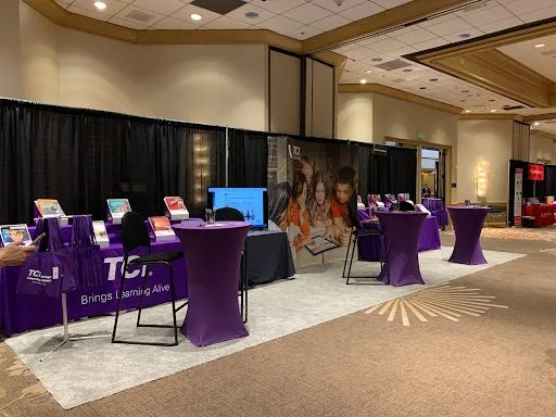 A convention booth with purple tablecloths, display screens, and learning materials arranged on tables.