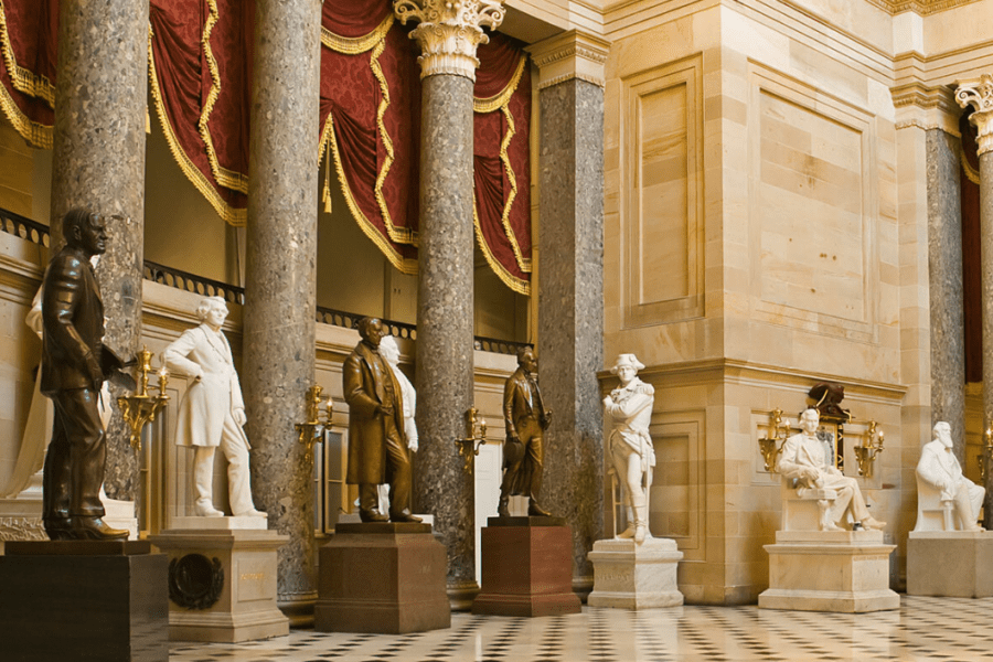 Marble and bronze statues line a grand hall with tall columns and red curtains in the background.