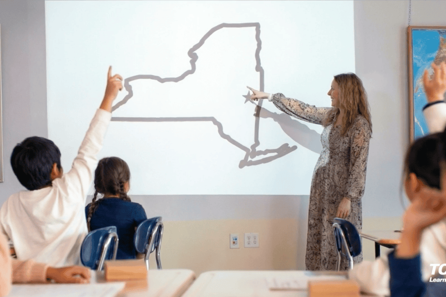 A teacher points to a map of New York on a projector while students raise their hands in a classroom.