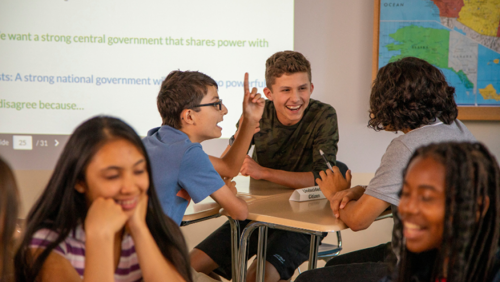 Students sit at desks in a classroom, talking and laughing together near a map and a projected screen.
