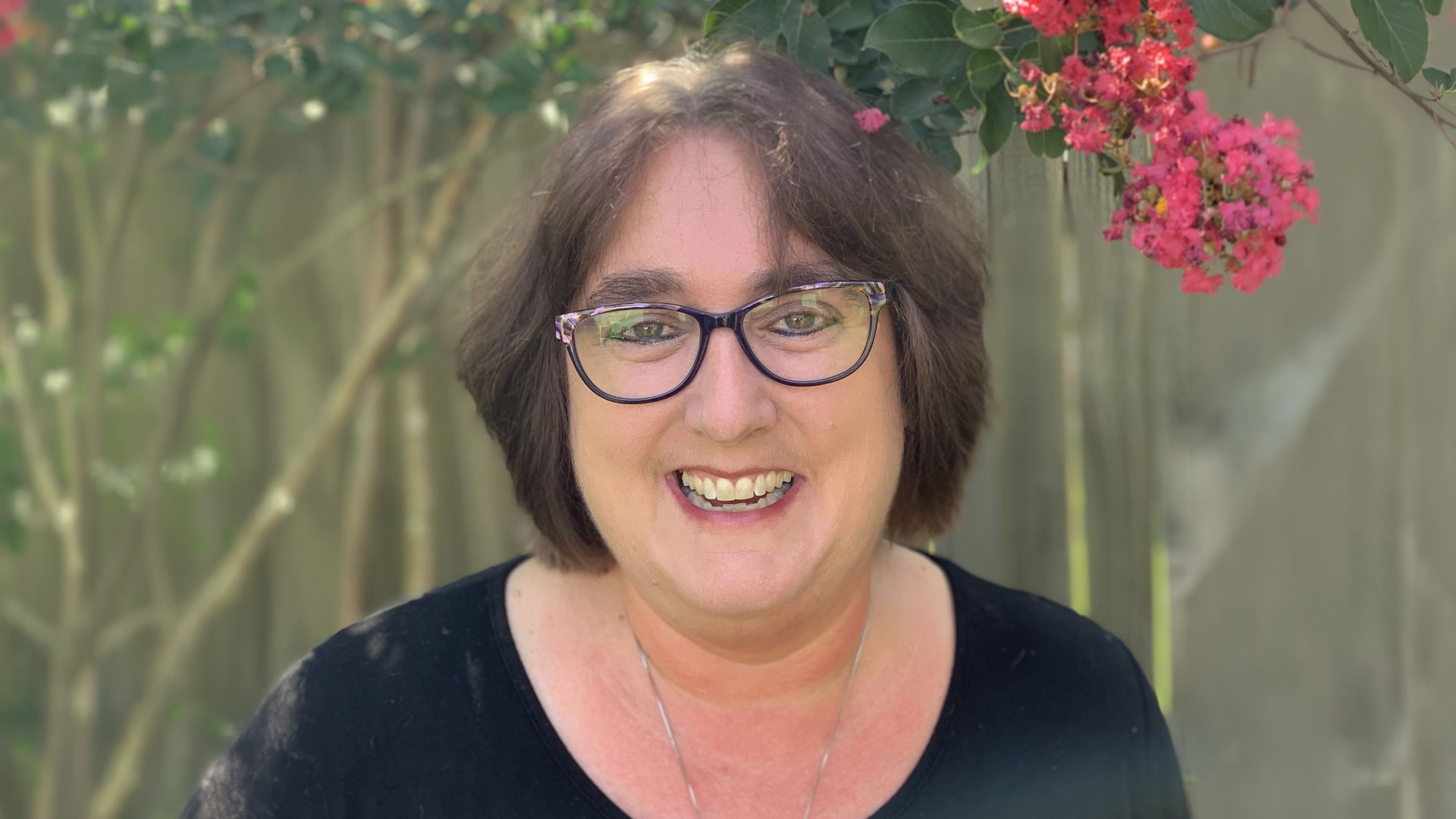 Woman with glasses smiling outdoors, standing in front of pink flowers and a wooden fence.